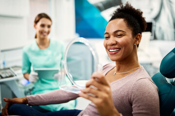 Happy black woman looking her teeth in a mirror after dental procedure at dentist's office.