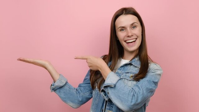 Young brunette woman 20s years old wears denim jacket pointing fingers hands aside on workspace copy space mockup promo commercial area, wears denim jacket, isolated on pastel pink background studio