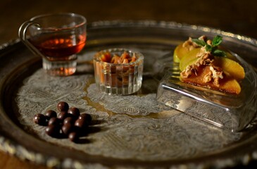 Small glass with liquor, nuts in a glass pot, peaches in syrup in small glass tray on a metal tray on a table.
