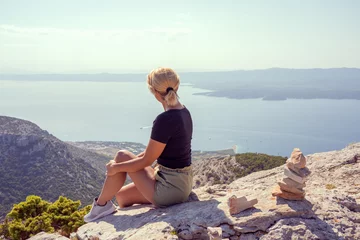Papier Peint photo autocollant Plage de la Corne d'Or, Brac, Croatie A girl sits on the edge of a cliff and looks at the sea. The girl looks from the mountain to the beach zlatni 
