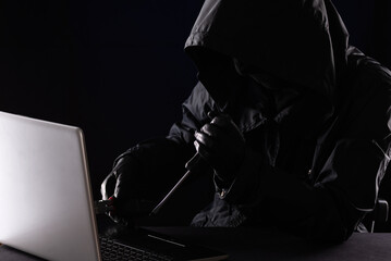 Criminal hacker in a hood in black clothes and balaclava destroys a laptop with tools, a screwdriver and pliers on a black background