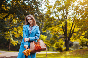 Portrait of young woman holding brown handbag in autumn park. Stylish girl wears trendy blue coat walking outdoors