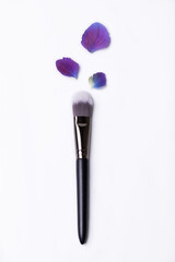 Makeup brush with white bristles isolated on a white background with purple and blue flower petals depicting natural beauty product.