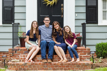 A family with a mother, father, and two daughters sitting outside on the brick porch of a small...