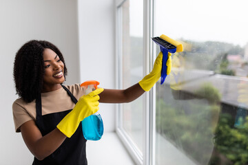 Beautiful young african woman is using a duster and a spray, looking at camera and smiling while...