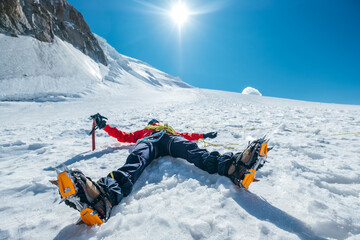 Tired exhausted climber lying under Mont Blanc du Tacul mountain. Wide opened legs in boots with crampons and mountaineering clothes with blue sky with bright sun background. Active people concept. - obrazy, fototapety, plakaty
