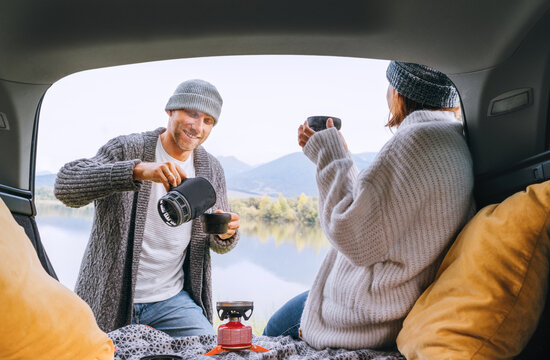 Car trunk view of chatting couple dressed warm knitted clothes enjoying gas stove prepared a drink with mountain lake view. Man pouring a fresh coffee. Cozy early autumn auto traveling concept image.