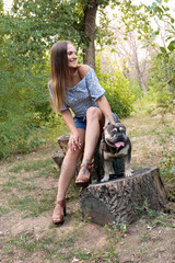 A girl and her dog French Bulldog are sitting on a tree stump in a park among green trees.