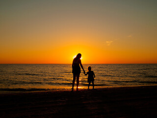 A little girl holds her mother's hand, standing on the sea beach on a summer evening. In the background, the evening sun setting over the horizon.