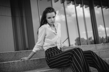 A melancholic black and white portrait of a young girl. A brunette in a white shirt and striped trousers is sitting on the steps