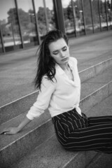 A melancholic black and white portrait of a young girl. A brunette in a white shirt and striped trousers is sitting on the steps