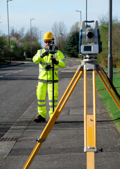 Land surveyor performing initial survey of the road levels and kerb lines before start of construction works using robotic tacheometer controlled by remote control and prism on pogo. - obrazy, fototapety, plakaty