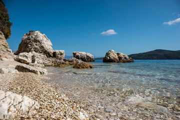 Fototapeta na wymiar White pebbles and rocks beach tide close-up with azure clear water on coast of Lefkada island in Greece. Summer vivid nature travel to Ionian Sea