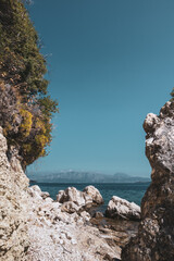 Rocky cliff with Mediterranean greenery and blue sky on pebbles beach on coast of Lefkada island in Greece. Summer wild nature travel to Ionian Sea. Vertical, color graded