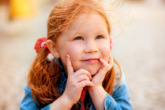 little smiling girl with red hair sitting on the road and thinking