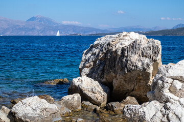 Big rocks in blue water on coast with white sail yacht and mountains on horizon, Lefkada island in Greece. Summer vivid nature travel to Ionian Sea