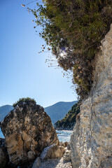 Sun shine through greenery on rocky cliff with blue sky on pebbles beach coast of Lefkada island in Greece. Summer wild nature travel to Ionian Sea. Vertical