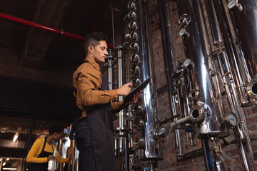 Caucasian man wearing apron holding a clipboard checking equipment at gin distillery