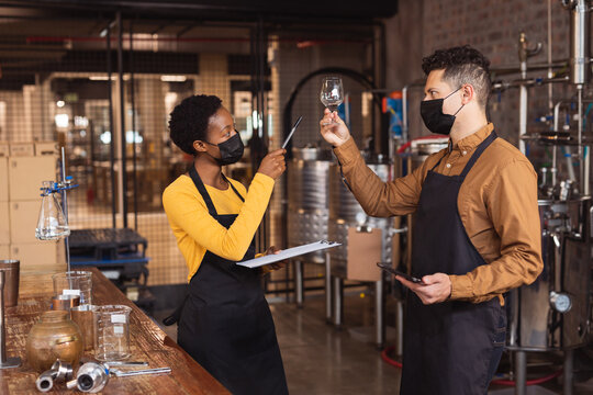 Diverse man and woman wearing face masks checking gin product in glass at gin distillery