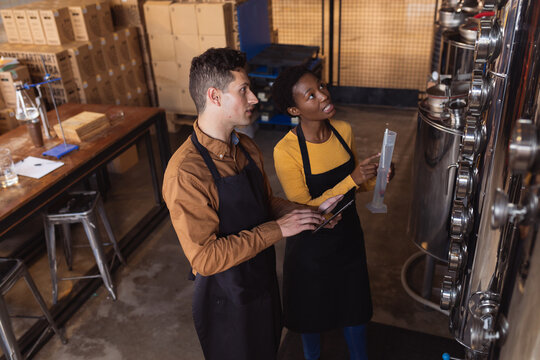 Diverse man and woman holding digital tablet and flask checking equipment at gin distillery
