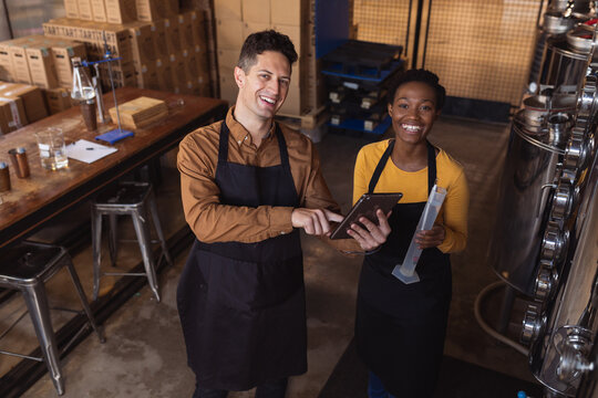 Portrait of diverse man and woman holding digital tablet and flask smiling at gin distillery