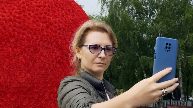 A beautiful woman in glasses makes a selfie on a smartphone in the park against the background of a composition of red roses. Special photo spots to attract visitors.
