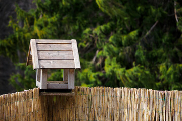 Wooden bird feeder on a railing with spruce in the background and reeds on the railing.