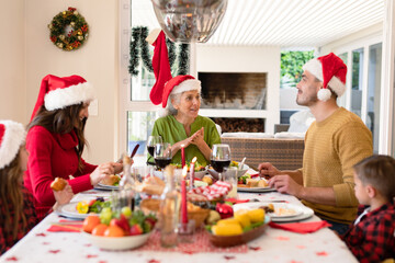 Happy caucasian multi generation family wearing santa hats sitting at christmas table