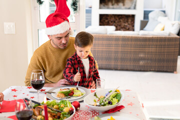 Happy caucasian father and son wearing santa hats sitting at christmas table