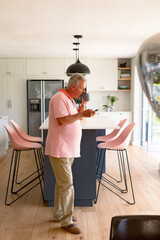 Relaxed caucasian senior man standing in kitchen, drinking coffee and using smartphone