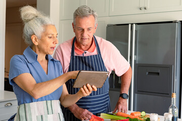 Happy caucasian senior couple standing in kitchen, using tablet and preparing meal together