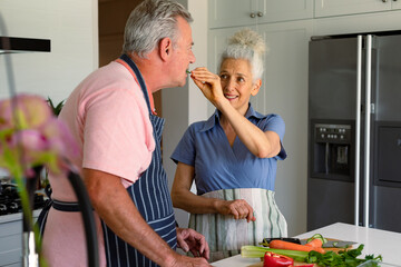 Obraz premium Happy caucasian senior couple standing in kitchen, preparing meal together and eating vegetables