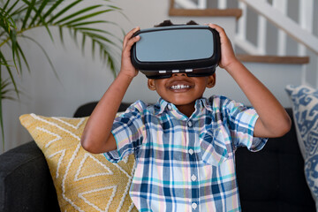 Happy african american boy sitting on sofa, using vr headset and having fun