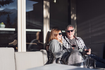 Happy middle aged couple sitting on an outdoor couch enjoying wine - 458636734
