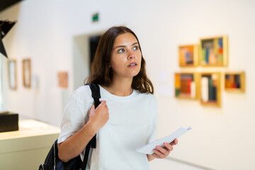 Thoughtful young girl visiting an art gallery exhibition of paintings studies the artwork with...
