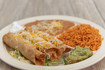 Taquitos rancheros, covered in shredded cheese and served on a plate with guacamole, rice, and...