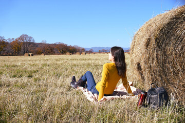 Naklejka na ściany i meble Woman in yellow sweater and rubber boots sitting on plaid next to backpack and thermos with hot tea near haystack in field. she looking thoughtfully into the distance mountains and the autumn forest.