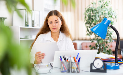 Concentrated young businesswoman working with laptop and papers at office desk..