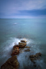 vertical seascape hot of rocky shore with stop motion of soft sea wave and blurred moving cloud with slow shutter in sun shine at morning scene