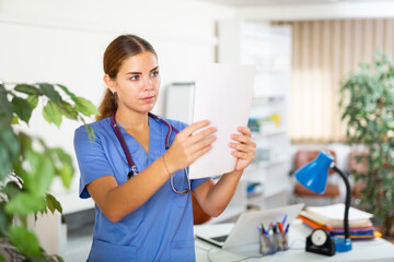 Portrait of young woman doctor standing in medical office with papers in hands, focused on studying of clinical diagnosis of patient