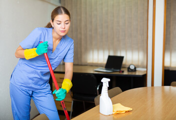 Portrait of professional female worker of office cleaning service wearing uniform and rubber gloves wiping floors with mop