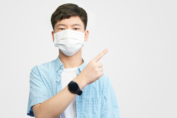 Portrait of a healthy smart asian teenager boy in casual blue with medical face mask looking to camera and pointing up. White background, New normal, Back to school, Poster, Copy space, Backdrop