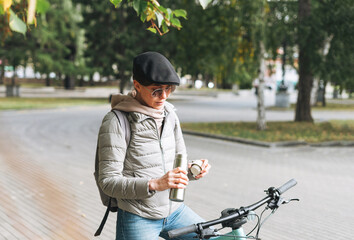 Portrait of fashionable young pretty woman in cap and sunglasses on bicycle with thermo mug on sunny autumn day in city park