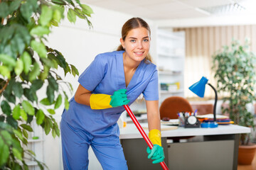 Woman in surgical scrubs and rubber gloves cleaning floor with mop in hospital.