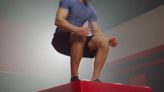 Young Caucasian fitness man in shirt and shorts jumping from the floor to the box with both feet, landing in squat position then step back to the floor and repeat, doing a box jump workout in a gym.