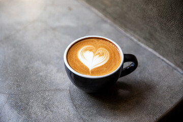 Black cup of cappuccino with latte art of heart shape on saucer on concrete background.