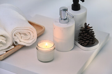 Burning wax candle, towels and shampoo bottles on white table.