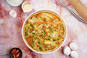 Baked french vegetable pie quiche filled with carrots, peas and cheese on a pink marble surface, cracked egg shell, flour, cream, top view