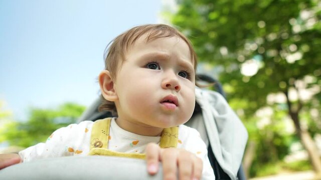 Curious little baby girl staring at other kids playing in the playground while she is sitting in a stroller with a serious expression, face close-up in slow motion, low angle