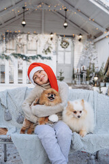 A little boy dressed in a red Santa hat holds two Spitz puppies in his arms. The child sits with dogs on the background of a white winter house. Christmas present for kid - 458667526